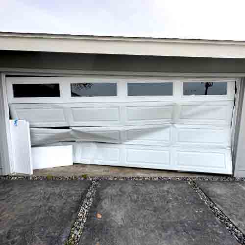 A photo of a garage door with a crushed panel and visible damage from a vehicle impact, showing the need for urgent structural assessment and repair.